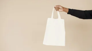 A hand holding up a plain white cotton tote against a beige background.