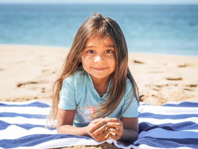 A young girl in a light blue short-sleeve swim top lying on a striped towel at the beach.