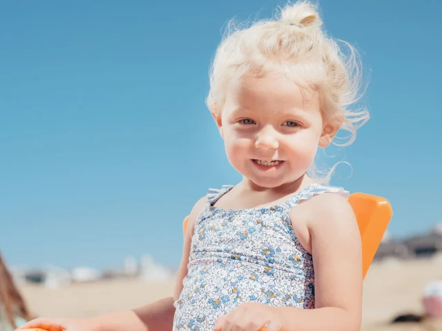 A toddler in a floral one-piece swimsuit with ruffled straps on a sunny beach.