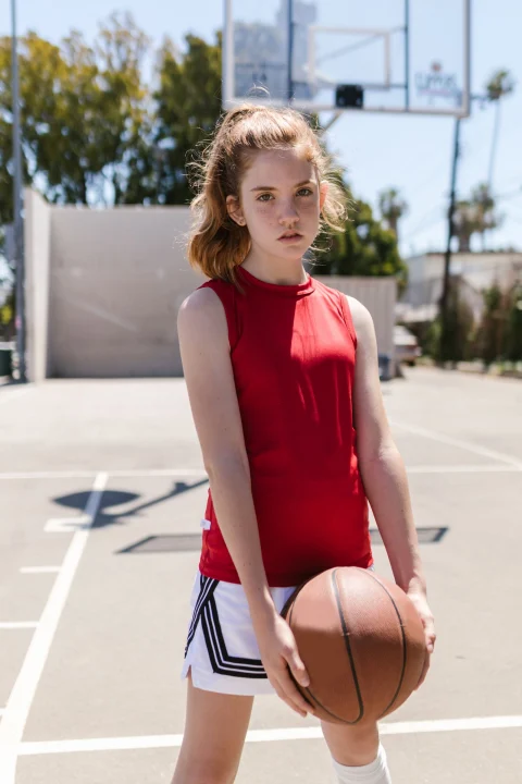 A girl in a red sleeveless top and white shorts with black side stripes, holding a basketball on an outdoor court.