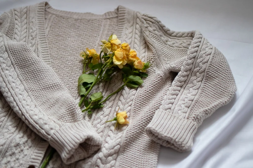 Oatmeal cable-knit cardigan laid open on white bedding with a few yellow roses on top.