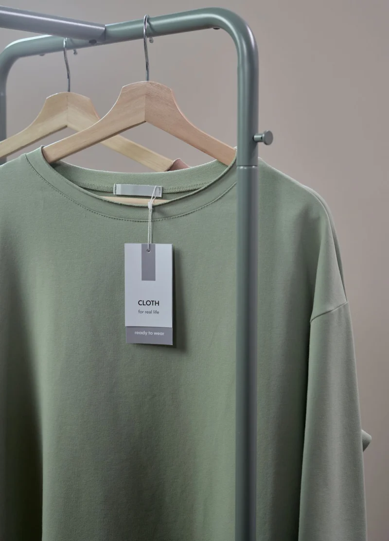 A sage green T-shirt on a hanger with a plain brand hang tag at the neck.